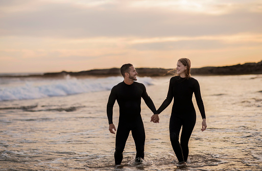 Happy Couple Holding Hands While Walking On The Beach Together, Romantic Surfers In Wetsuits Standing In Water, Relaxing After Surfing, Enjoyijg Spending Time With Each Other, Copy Space