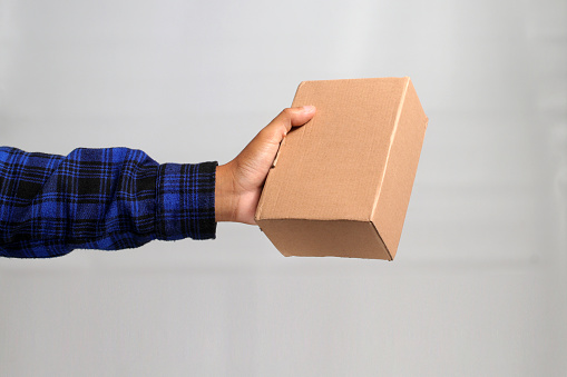 A hand holding a cardboard box against a gray background, illustrating the concept of shipping, delivery, and logistics services