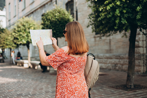 Happy Tourist girl with paper map walking in the city during vacation. Cheerful 30s woman in sunglasses traveling abroad in summer. Travel and active lifestyle concept
