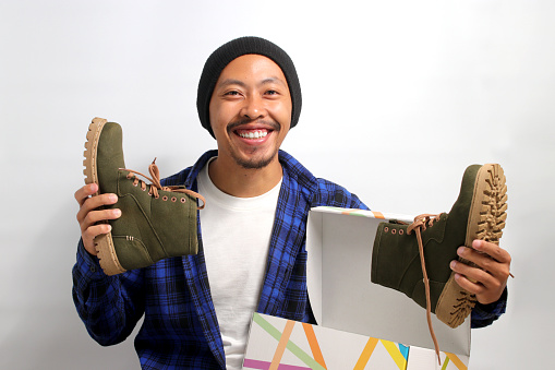 Happy Asian man, sporting beanie hat and casual shirt, joyfully unboxes a parcel package containing new shoes against a white background. Online business influencer showcasing products on social media