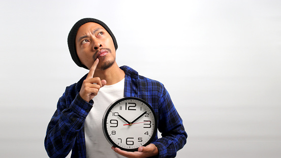 Pensive Asian man, dressed in a beanie hat and casual shirt, gazes up at empty copy space, deeply lost in thought, while holding a clock and resting his hand on his chin, Isolated on white background