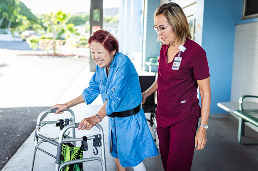 A caring Eurasian mature adult occupational therapist helps her female elderly patient walk safely with the assistance of a transfer gait belt as they meet and work together outside the patient's apartment complex.