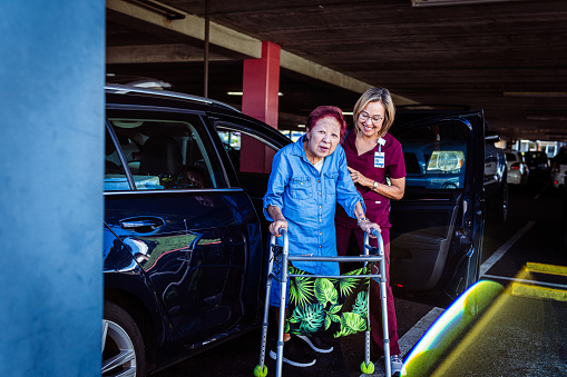 A kind mature adult Eurasian healthcare worker wearing scrubs helps her elderly patient who is using a mobility walker as they walk together through a parking garage.