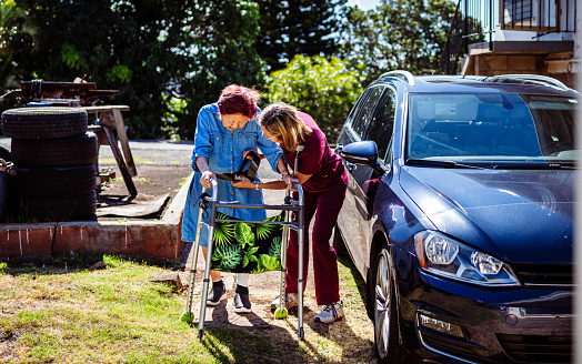 A caring female nurse helps an elderly woman of Asian ethnicity use a walker to walk across a residential driveway.