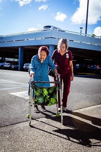 A kind mature adult nurse of Hawaiian and Finnish descent walks with her elderly patient who is using a mobility walker through a parking lot on a sunny day, with a parking garage visible in the background.