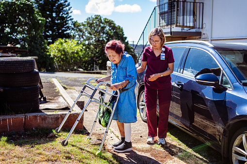 A caring female nurse helps an elderly woman of Asian ethnicity use a walker to walk across a residential driveway.