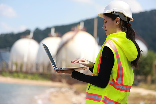 Beautiful engineer lady standing in front of chemical tanks in a special uniform. Female engineer uses a laptop to work outside.