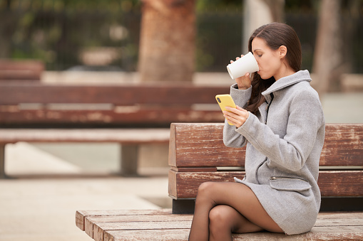 Young woman drinking coffee sitting on a brown bench while checking her smartphone during her break time.