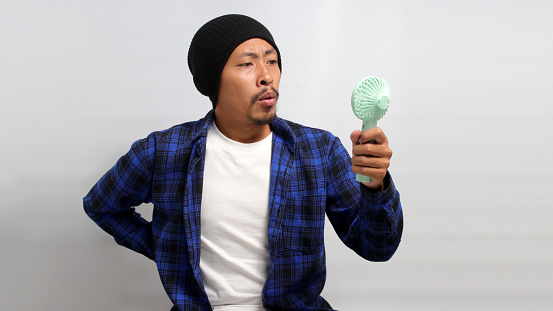 Sweaty young Asian man, dressed in a beanie hat and casual shirt, feels overheated and is visibly suffering from the summer heat. He uses handheld portable mini fan in an attempt to cool himself down