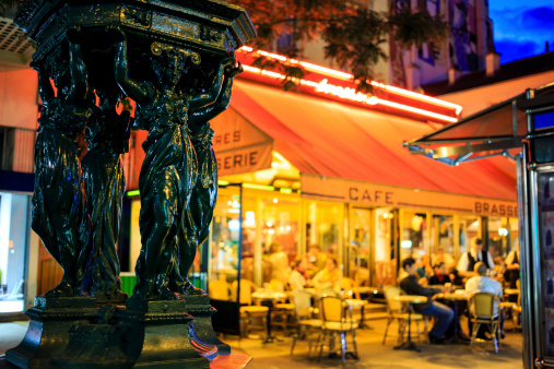 Paris by night - antique drinking fountain with brightly lit street café on the background. Montparnasse district, Paris