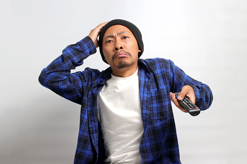 Unhappy Asian man, dressed in beanie hat and casual shirt, touches his head while holding a TV remote control, feeling disappointed and sad as he watches his favorite team lose in a sports game match
