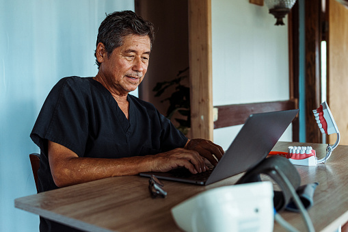 A mature adult male dentist of Hawaiian descent sits at a desk in his home office and uses a laptop computer to consult a patient virtually.