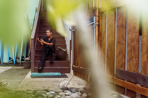 A mature adult multiracial man of Hawaiian and Finnish descent who is wearing scrubs sits on the stairs in front of his house in Hawaii and sends a text on his smart phone after returning home from work.
