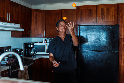 An active and healthy senior man of Hawaiian and Finnish descent who just arrived back home from work and is wearing black scrubs stands in the kitchen of his home and smiles while juggling oranges.