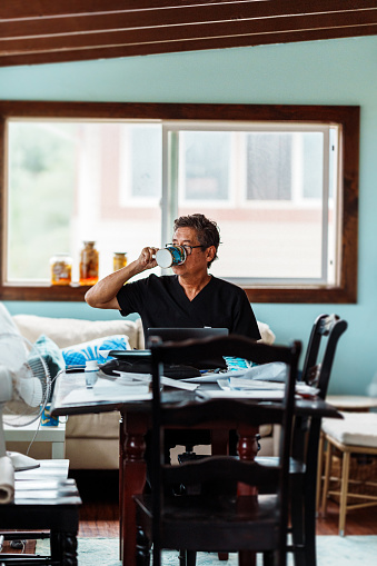 A active senior man of Hawaiian and Finnish descent who is wearing scrubs takes a sip from a mug while looking through mail and working on a laptop computer in the dining room of his home.