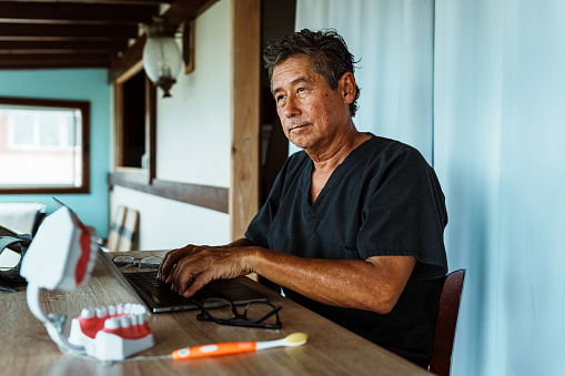 A active and healthy senior male dentist of Hawaiian and Finnish descent who is wearing scrubs sits at a desk in his home office and works on a laptop computer.