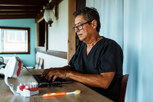 A active and healthy senior male dentist of Hawaiian and Finnish descent who is wearing scrubs sits at a desk in his home office and works on a laptop computer.