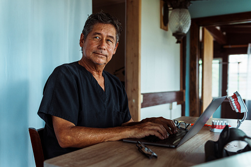 A handsome senior male dentist of Hawaiian and Finnish descent sits in his home office and looks off into the distance with a contemplative expression.