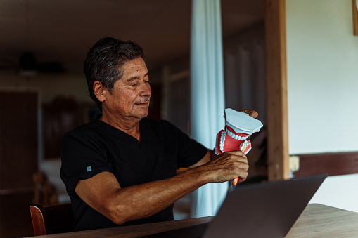 A handsome mature adult dentist of Hawaiian descent holds a model of teeth while consulting a patient virtually using telemedicine health technology from his home office.