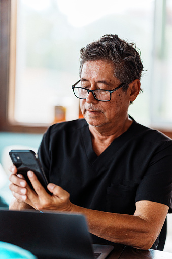 A male medical profession of Asian descent glances down at his cell phone while working on a laptop computer in a business office.