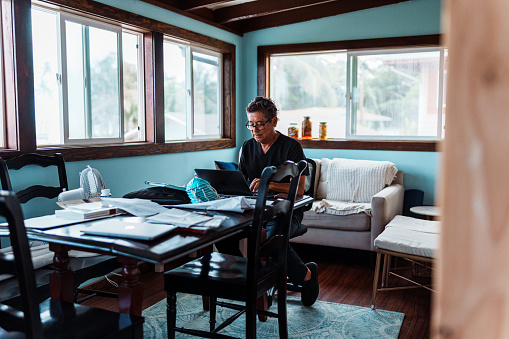 A active senior man of Hawaiian and Finnish descent who is wearing scrubs looks through paperwork and works on a laptop computer in the dining room of his home.