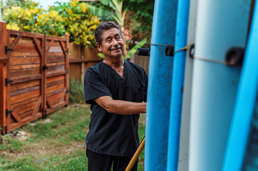 A vibrant mature adult multiracial man of Hawaiian and Finnish descent who is wearing scrubs stands outside his house and chooses a surfboard to load into his car before heading to work in the morning.