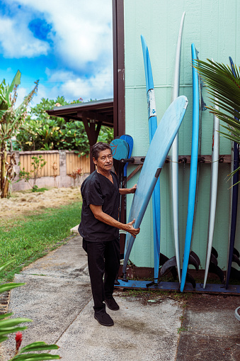 A vibrant mature adult multiracial man of Hawaiian and Finnish descent who is wearing scrubs stands outside his house and chooses a surfboard to load into his car before heading to work in the morning.