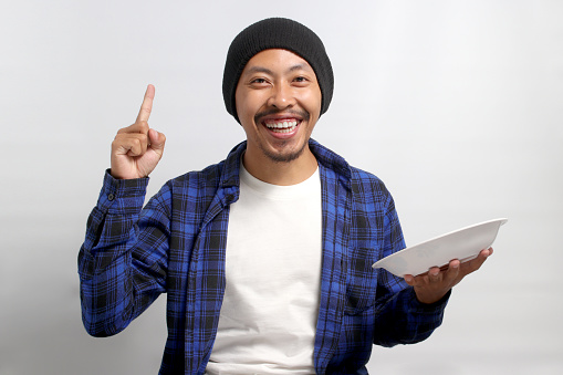 Young Asian man, wearing a beanie hat and casual shirt, raises his finger in a eureka moment while holding an empty white plate, smiling at the camera, and standing against a white background