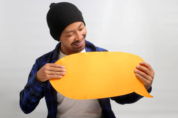 Excited young Asian man, dressed in a beanie hat and casual shirt, holds an empty speech bubble while standing against a white background