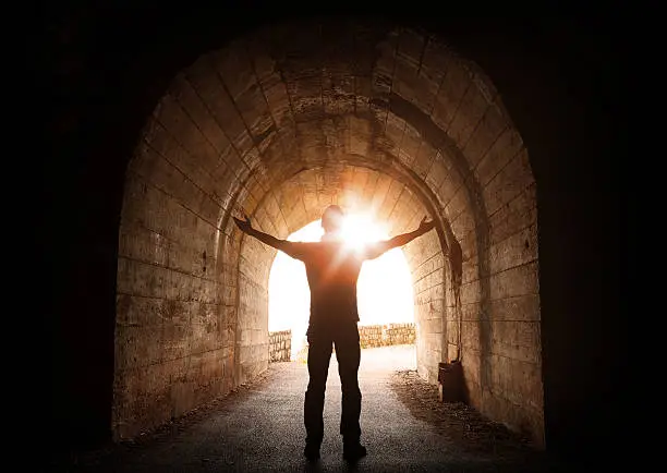 Man stands inside of old dark tunnel with shining sun in the end
