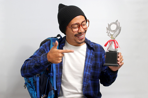 A young Asian male student, dressed in eyeglasses, a beanie hat, and a casual shirt, carries a backpack while pointing at his winning trophy, while standing against white background