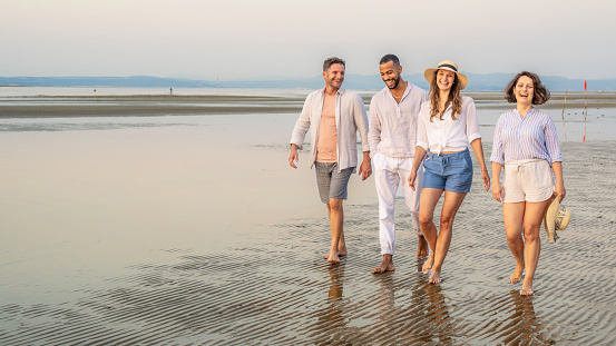 Diverse group of friends laughing while walking along coastline barefoot and looking at camera, wide shot portrait