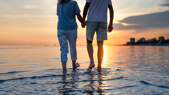 Senior adult couple silhouettes holding hands and walking to sea barefoot at sunset, low section back view