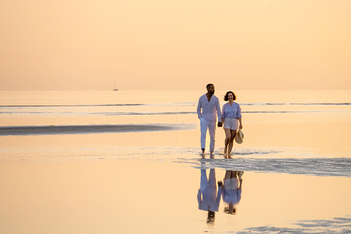 Couple holding hands and walking along coastline at golden sunset, wide shot