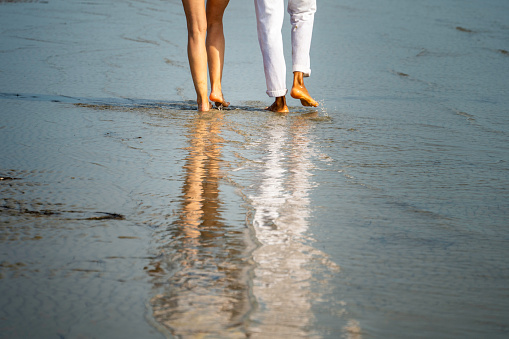 Man and woman couple walking along coastline barefoot, low section back view view