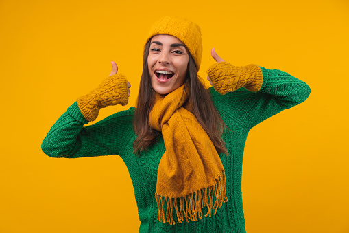 Studio portrait of attractive young woman with charming smile posing in cozy knitted sweater, beanie hat, scarf and mittens over bright yellow background showing thumbs up gesture.