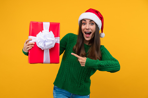 Studio shot of beautiful happy joyful excited girl wearing Santa hat and green knitted sweater is pointing with index finger at decorated gift box in her hand.