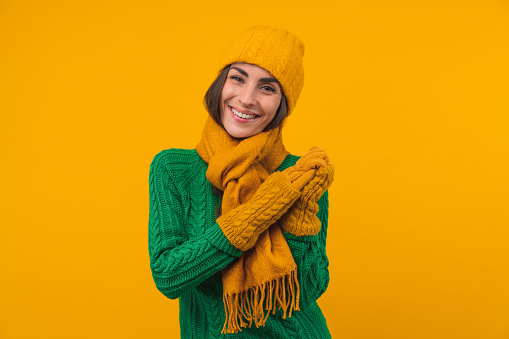Studio shot of beautiful young woman with charming smile posing in cozy knitted sweater, beanie hat, scarf and mittens isolated over bright yellow background.