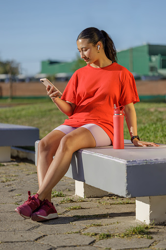Latin girl with wireless headphones using a mobile phone on a bench in a public park.