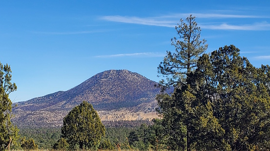 This photo captures the stunning view of Maroon Crater, an extinct volcano in Arizona. The sky is a brilliant blue with wispy clouds, and the desert landscape is dotted with rocks, shrubs, and grass. The volcanic cinder cone rises in the background, showcasing the geology and history of this scenic area.