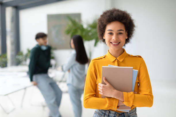 istock Cheerful black lady student posing with notepads and smiling at camera while her classmates talking on background 1863487294