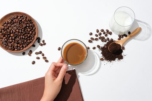 Hand holding a cup of coffee, coffee beans on a wooden plate, a glass of milk, coffee powder and wooden spoon on a white background. Ideal space for advertising.