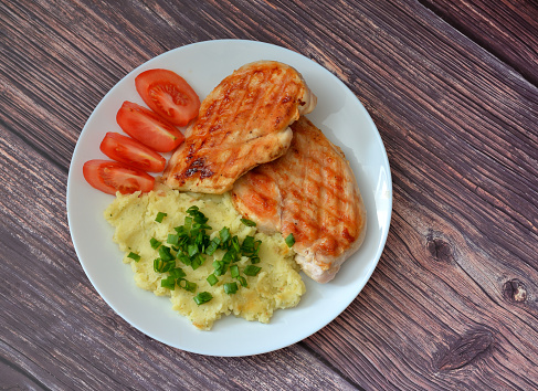 Two grilled chicken fillets with tomatoes and mashed potatoes with green onions on a wooden table. Top view, flat lay.