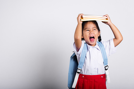 Asian adorable toddler smiling happy wear student thai uniform red skirt stand holding book over head and screaming in studio shot isolated on white background, Portrait little children girl preschool