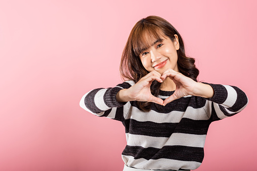 Portrait of a happy Asian woman with a confident smile, making a heart symbol with her fingers and hands on a pink background. Sending love and joy for Valentine's Day.