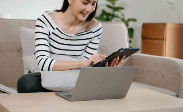 istock Young asian woman using smartphone and tablet while seated on couch at home 1864059712