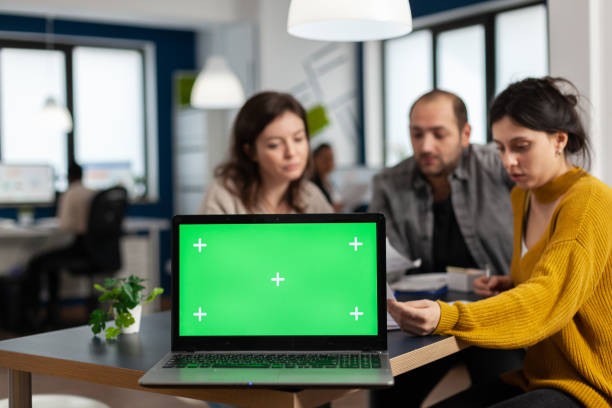 Laptop with green screen placed on desk while people work in background