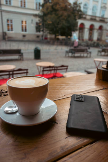 Coffee cup and mobile phone on wooden table near window. Sidewalk cafe, view from inside. Coffee break in cafe, Lviv, Ukraine. Morning coffee concept. Urban lifestyle.