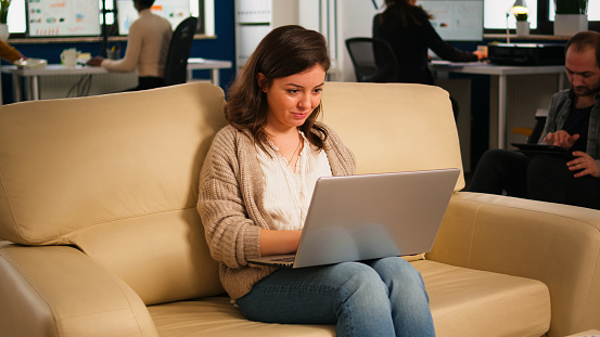 Manager woman searching on laptop sitting in relax zone on confortable couch typing on pc smiling while diverse colleagues working in background. Multiethnic coworkers planning new financial project