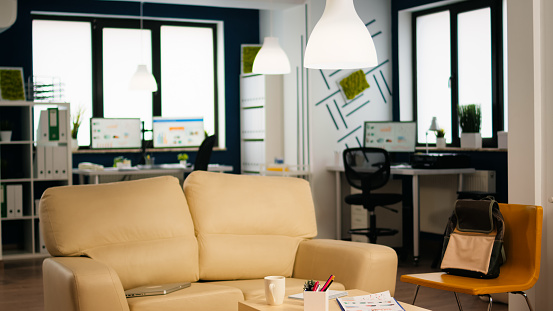 Interior of cozy light company room with small table and confortable couch, modern stylish chairs and desktop computer, all ready for employees. Empty spacious office of creative working space.